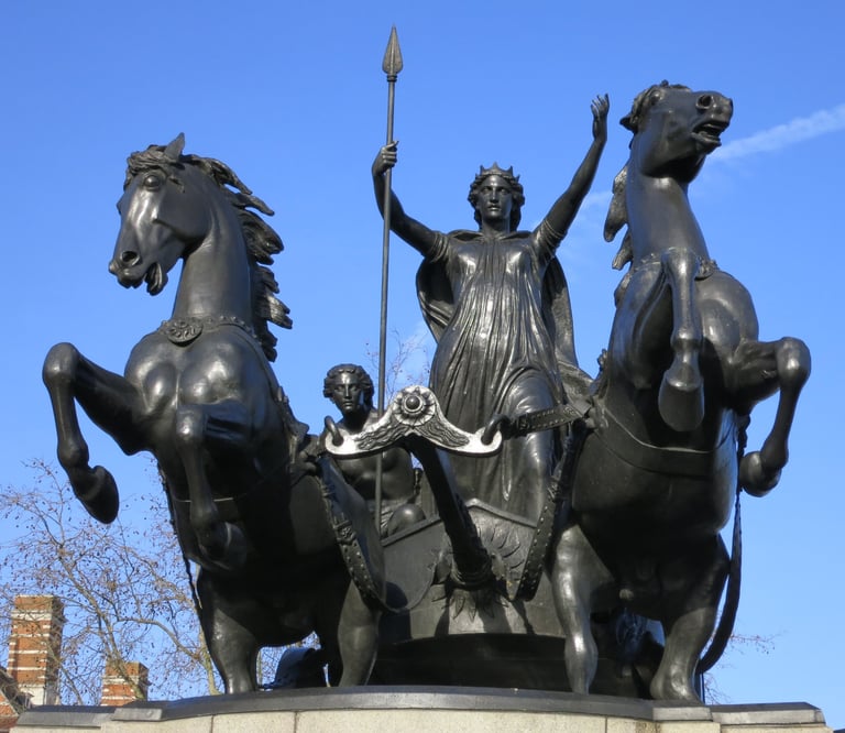 Bronze statue of a classical figure with raised arms holding a spear, flanked by two rearing horses against a blue sky